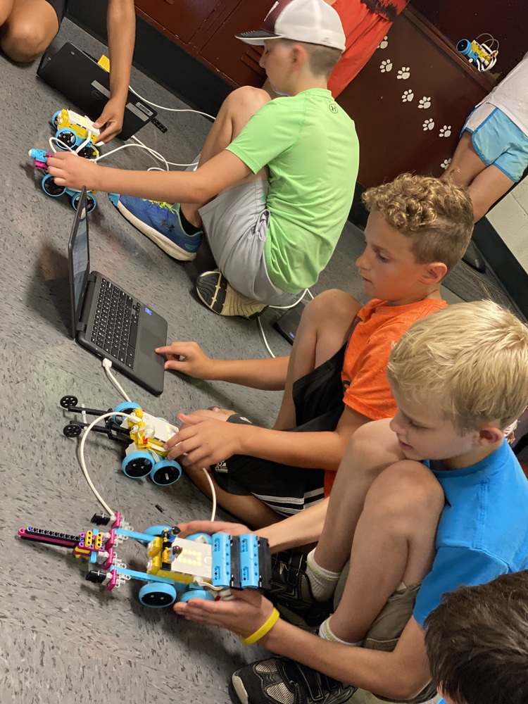 Three boys work on lego robotic cars they have made and programmed .  On has a laptop computer to use  in programming and operating the robots.