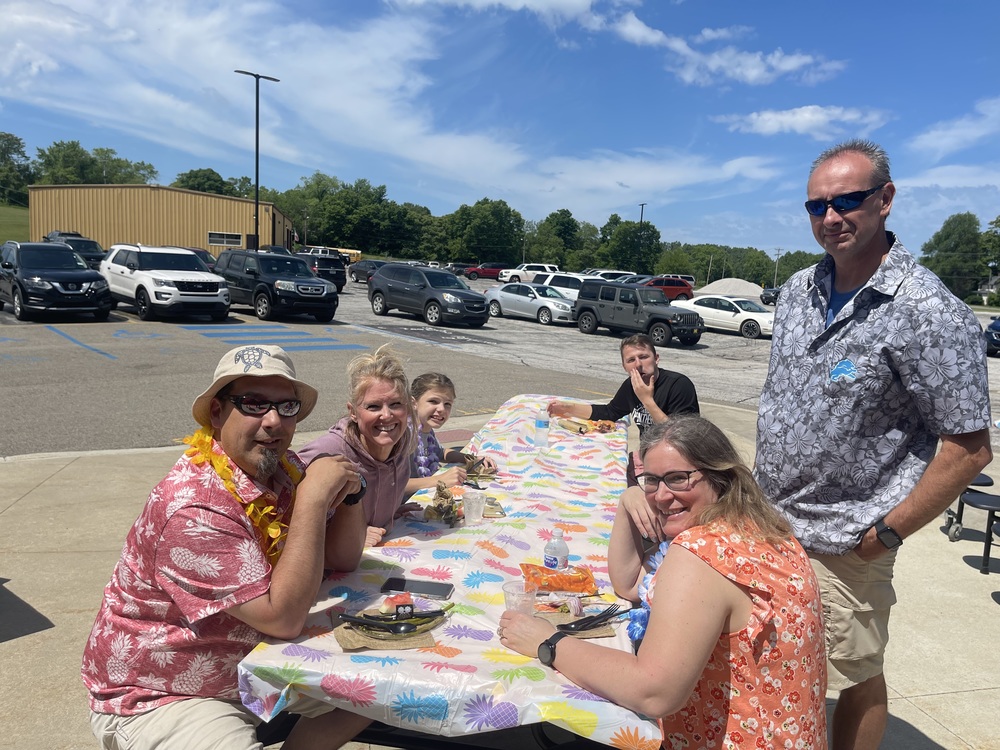 A group of educators wearing Hawaiian Shirts sit outdoors at a picnic table. One man is standing. The table is covered with a tropical print tablecloth.
