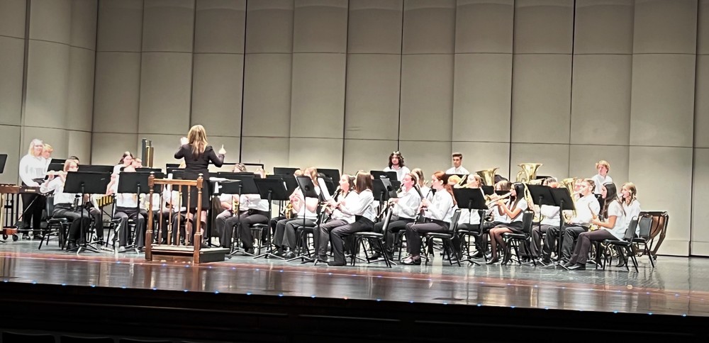 A female band conductor stands with back to the camera, as she conducts the band.  Band members are seated facing the camera playing instruments