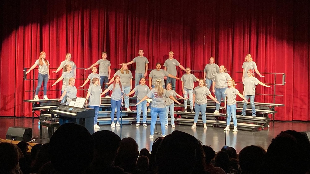 Watervliet High School Choir Students are standing on risers on the auditorium stage and performing.  They are all wearing jeans and grey t-shirts.