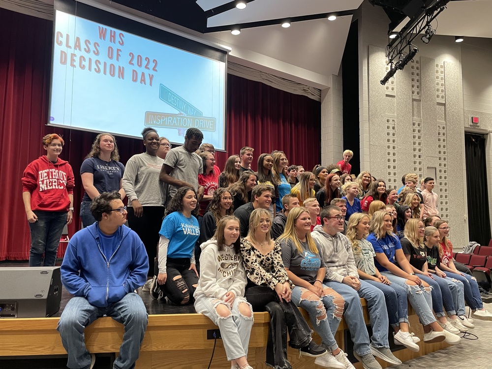 a large group of high school students sits and stands on the stage in the auditorium.  The screen behind them says "WHS Class of 2022 Decision Day"