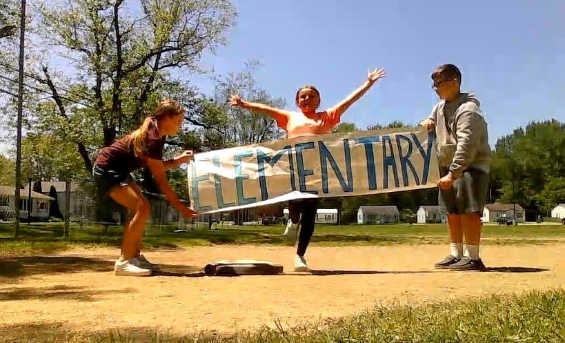 Two students hold a banner with the word "elementary" written on it in big blue letters.  A female student is running at the banner to run through it.