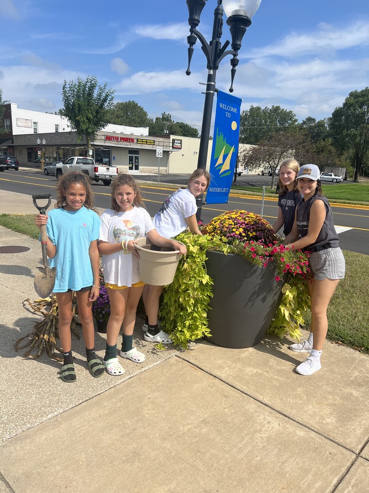 Five middle school age girls stand next to a large pot where they are planting mums in downtown Watervliet