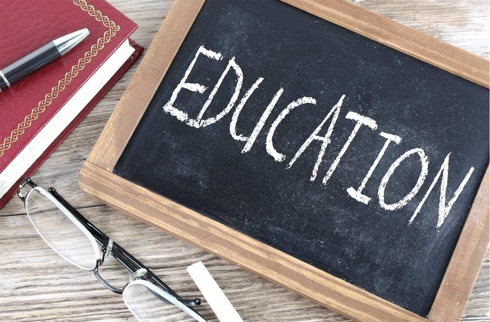 a chalkboard with the word "EDUCATION" written on it.  a pair of reading glasses and chalk  lie on the woooden table which holds the chalk board, and also a red bound-book and an ink pen,