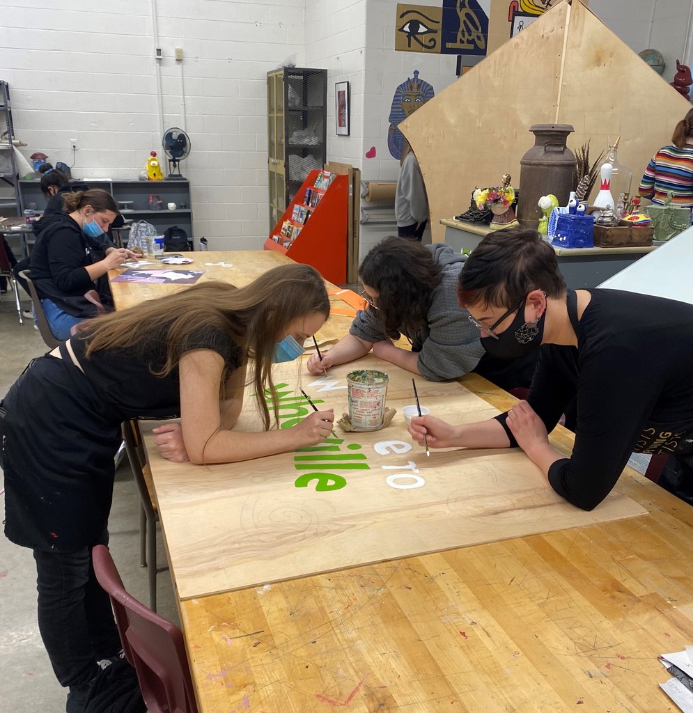 Female students and a teacher are painting a sign that says "Welcome to Who-ville" to be hung at the Watervliet Hometown Christmas Celebration.