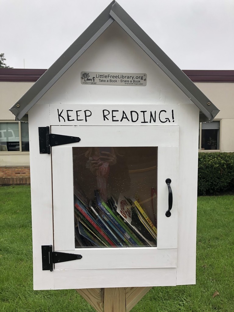 Photo of a house shaped wooden box with a door on the front.  There is a window in the door and you see books inside.  Text on the box:  Little Free Library.org, Take a Book-Share a Book.  Keep Reading!