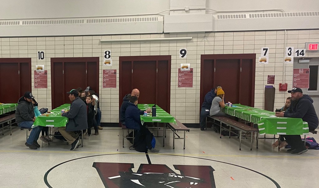Men and elementary school students are seated at long cafeteria tables  with green and white table coverings eating donuts at the Donuts for Dudes event.