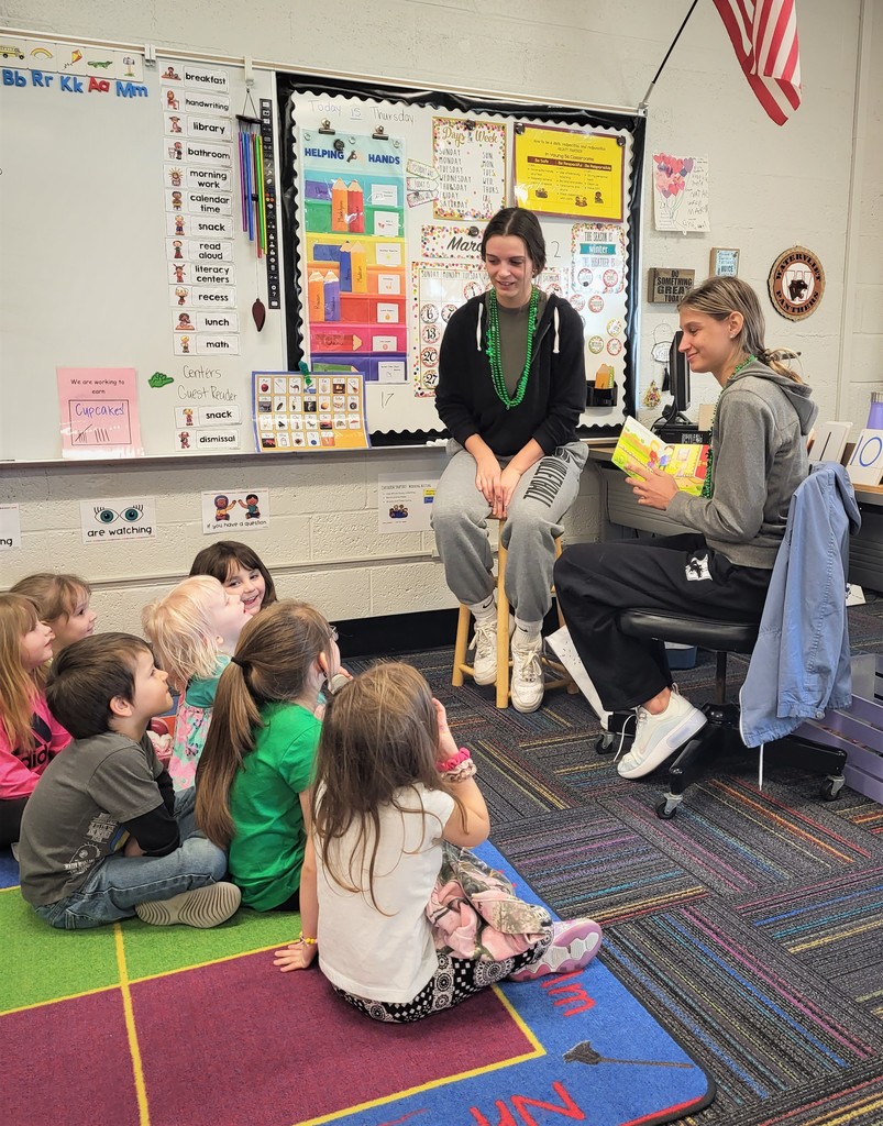 a group of young children sit on the floor in an elementary classroom listening as a high school age girl sits on a chair reading a story to them.  Another high school girl looks on.
