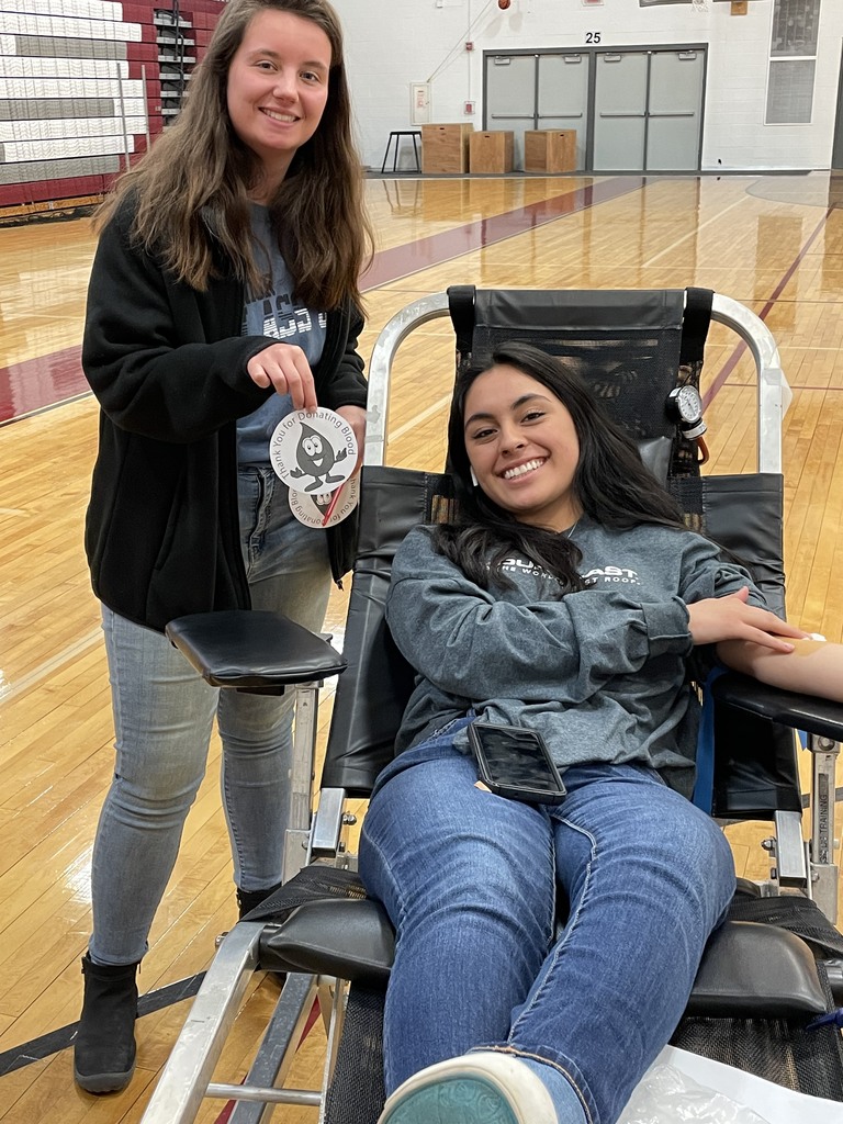 A high school girl is donating blood.  Another girl is holding a round cutout of a blood droplet with a smiling face that says Thank you for donating blood