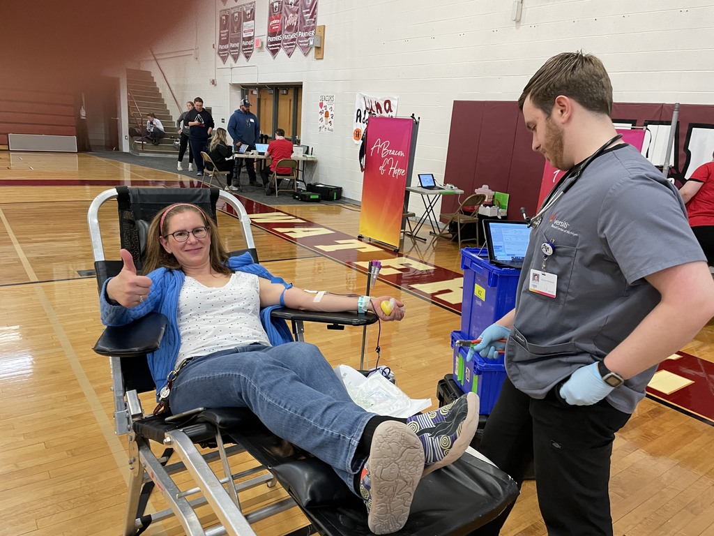 A female teacher is giving a thumbs up sign as she is giving blood