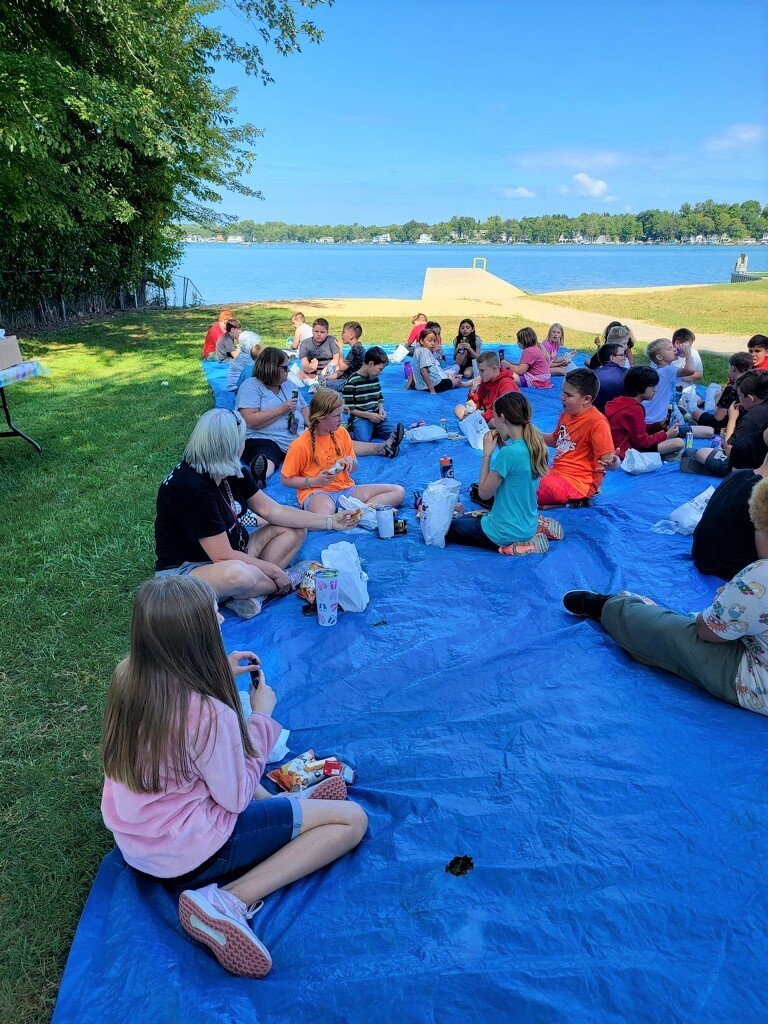 Students sit on a blue plastic tarp which is spread out on the lawn eating lunches.  Paw Paw Lake is in the background.
