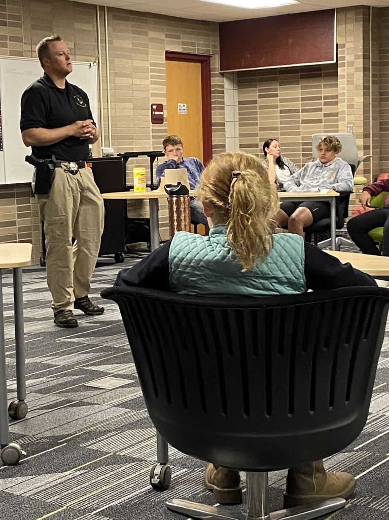 Sherrif Deputy Hahn is speaking to a group of middle school students.  The deputy stands at the front of the room and students are seated around him in chairs at tables.