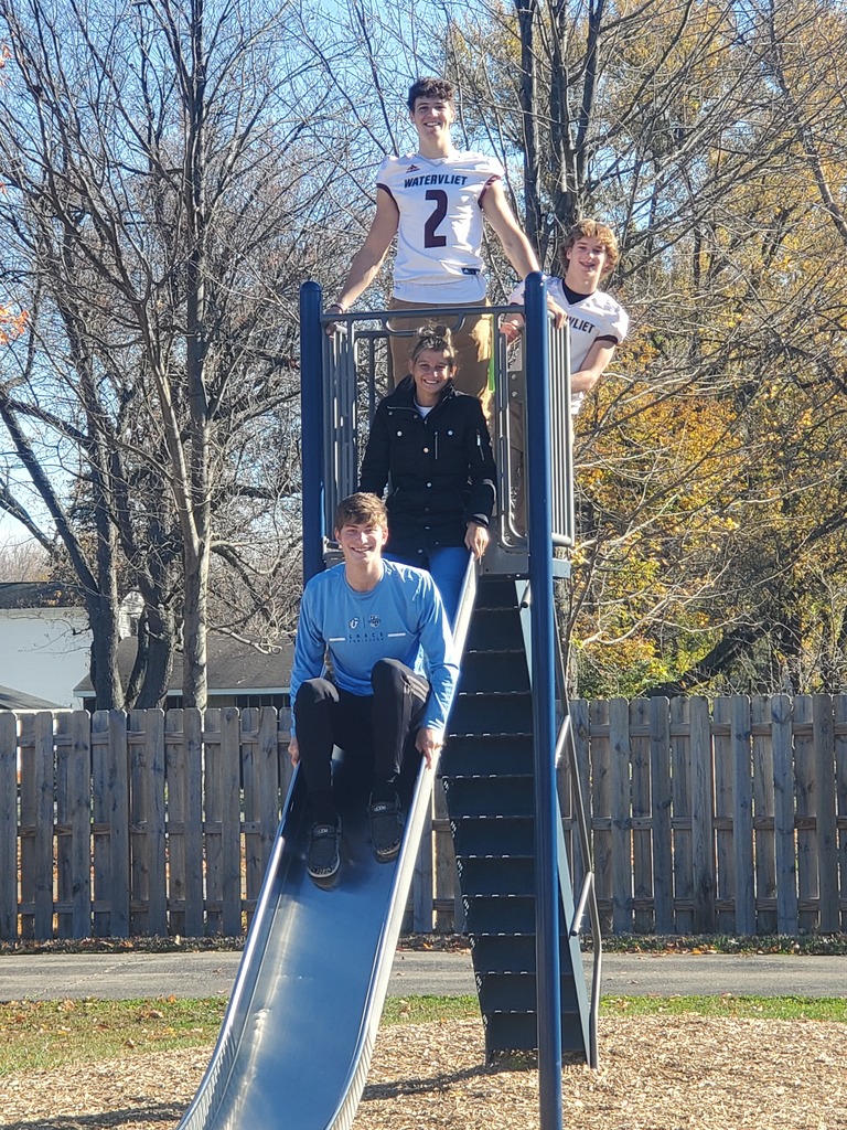 Three high school boys and one girl are pictured on the slide at North School playground. There are trees and a wooden fence in the background.