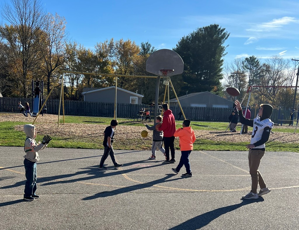 Some elementary students are playing basketball with a high school boy.  In the foreground, a young boy and a high school boy are tossing a football to each other.