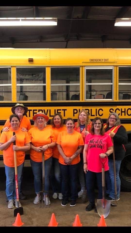 WPS School Bus drivers, most wearing safety orange T-shirts and holding rakes, shovels, or other garden tools, stand posed in front of the side of a school bus