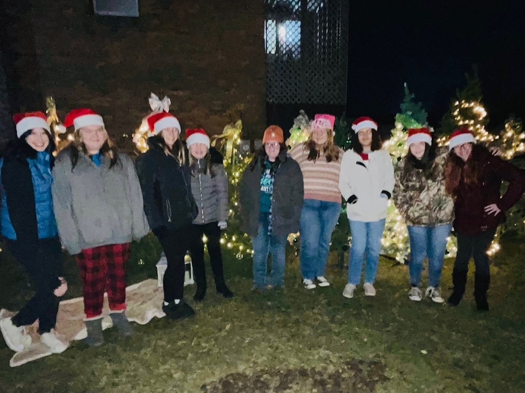 Nine choir students wearing Santa caps and winter coats are lined up in front of some Christmas trees outdoors