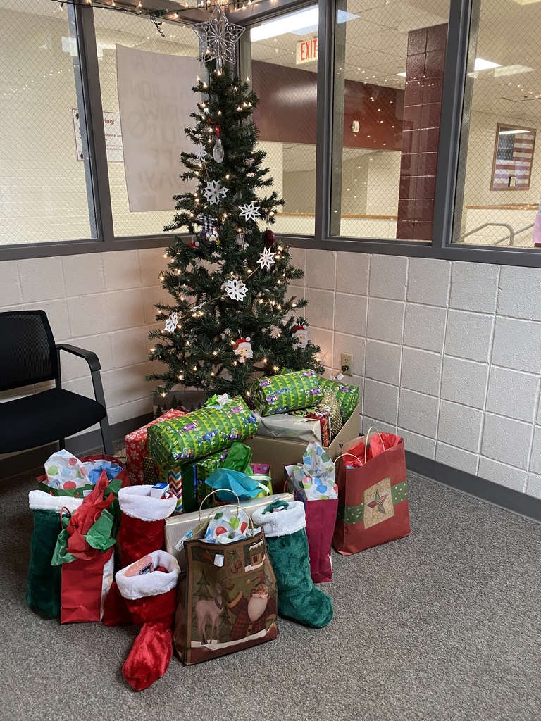 A christmas tree sits in the corner of an office with lots of wrapped gifts in front of it