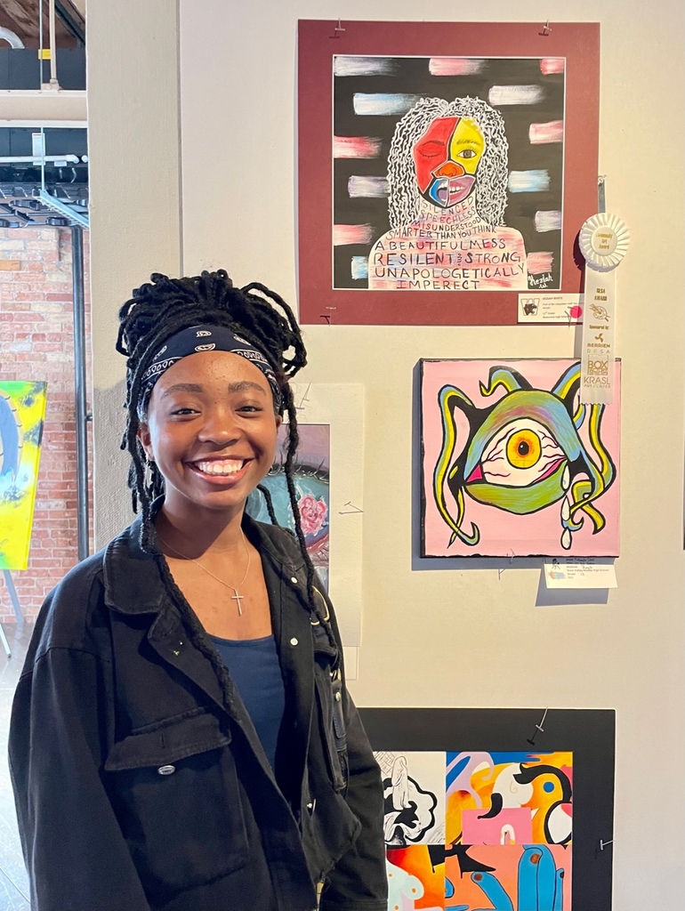 WHS Student Keziah White stands facing the camera smiling next to a wall of colorful artwork