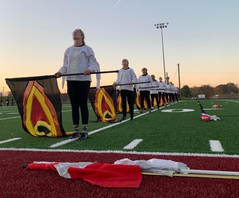 members of the band's flag section hold their flags horizontally at waist height.  They are standing on a yard line on the football field.